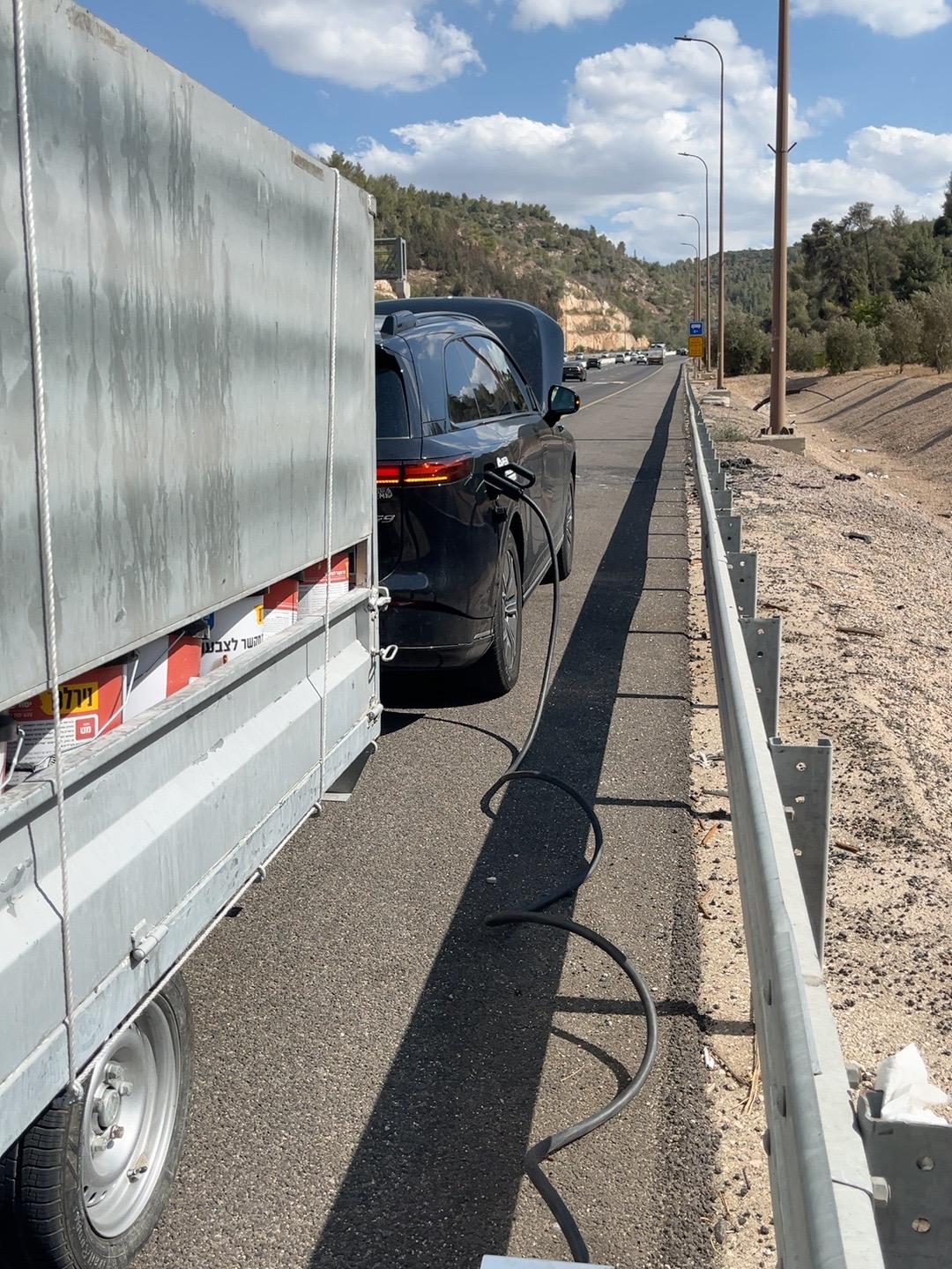 רכב חשמלי מחובר לכבל טעינה לצד ניידת שרון שירותי דרך וחילוץ בשולי הכביש